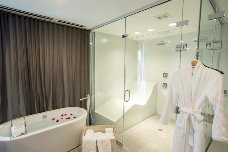A modern spa bathroom featuring a glass-enclosed steam shower with a built-in seating area, a freestanding bathtub with rose petals, and a hanging white bathrobe. Towels are neatly folded in a basket nearby, and a dark curtain separates the space for privacy.