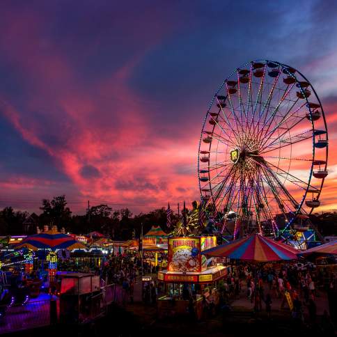 Great Frederick Fair in Frederick, Maryland, featuring a brightly illuminated Ferris wheel towering above carnival rides and vendor tents at sunset, with colorful lights and dramatic pink and purple skies creating a vibrant fairground scene.