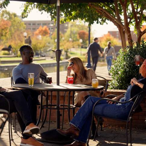Group seated at outdoor tables on the Idiom Brewery patio in Frederick, Maryland, with drinks on the table, metal chairs under umbrellas, trees and brick planters nearby, and a walkway and water feature visible in the background.