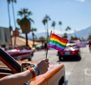 Pride flag waving outside of car in downtown Palm Springs with mountains and palm tree