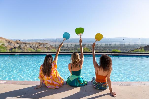 Three girl friends holding up fans and posing for a photo poolside at The Ritz-Carlton, Rancho Mirage.