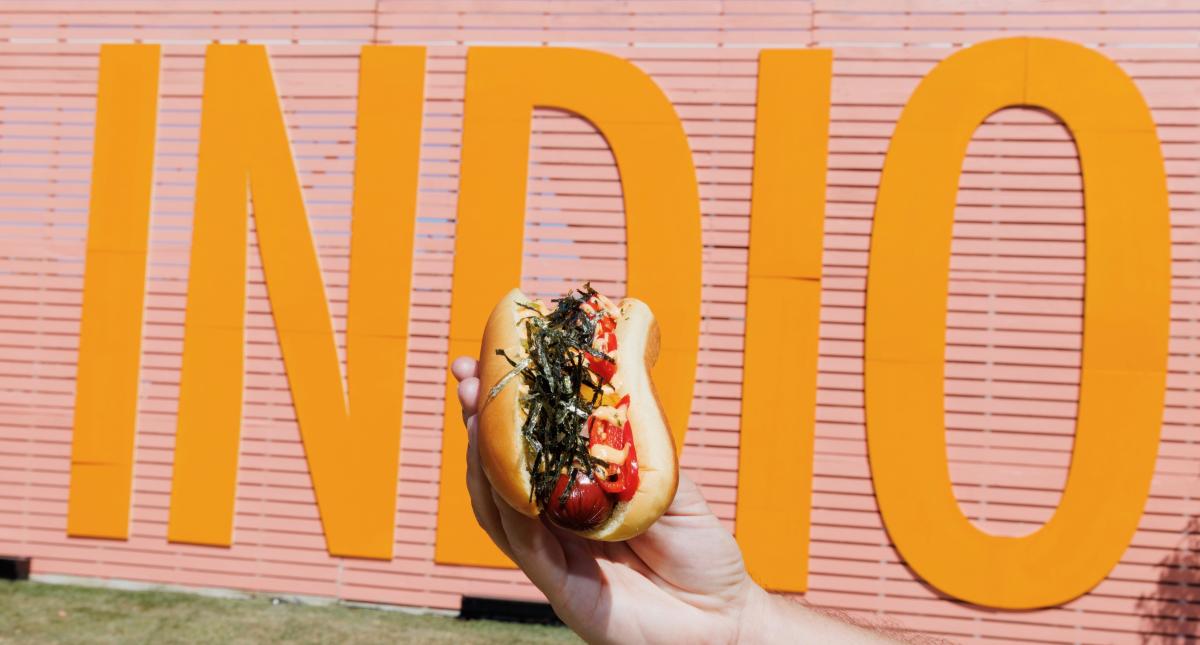 A person holds a gourmet hot dog in front of the Indio Central Market sign at Coachella festival.