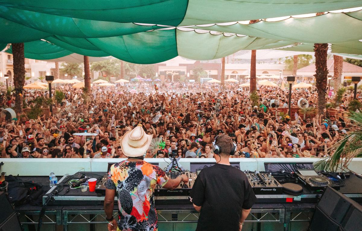 The backside of two DJs in a DJ booth with a horde of people in front of them underneath a green shade sail.
