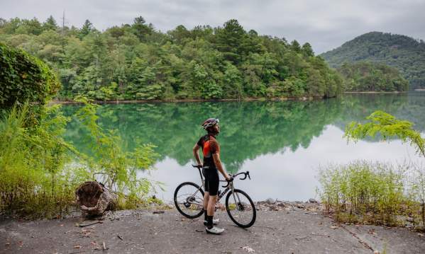 Man wearing helmet and cycling gear with a bike in front of a lake.