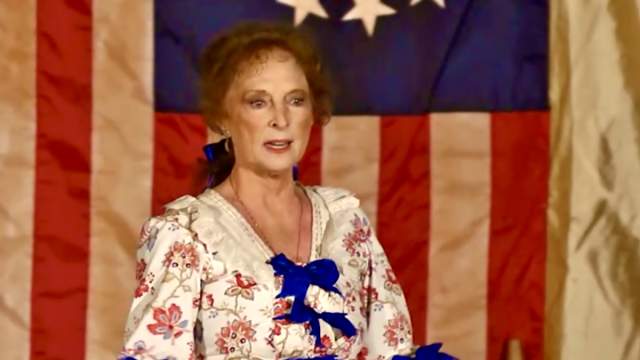A woman in a floral dress with blue bows stands before a colonial-era American flag.