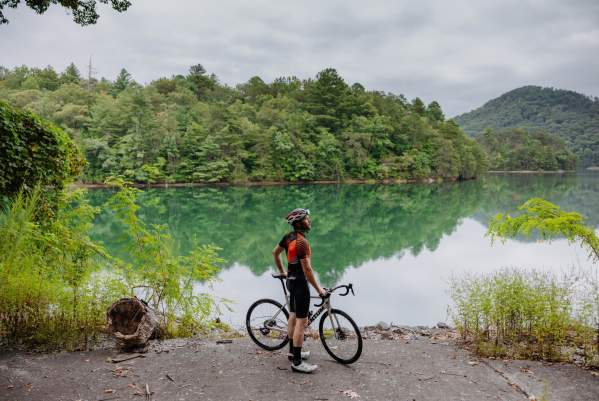 Man wearing helmet and cycling gear with a bike in front of a lake.