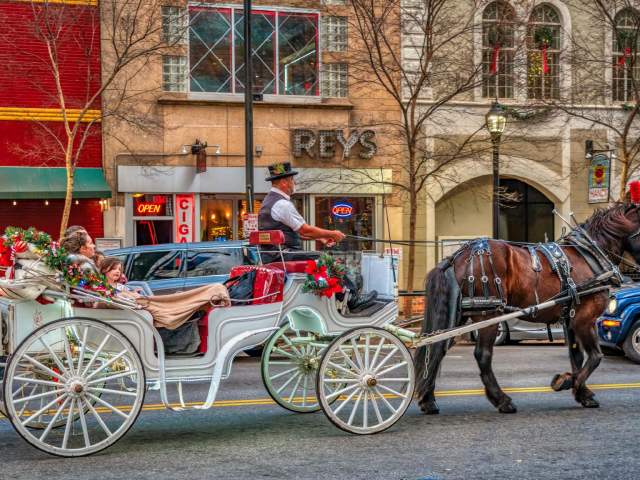 A white horse-drawn carriage decorated with Christmas garland and lights trots down Main Street in downtown Greenville, SC.