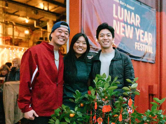 Three smiling people stand behind a potted kumquat tree decorated with red ornaments in front of a Lunar New Year Festival banner.