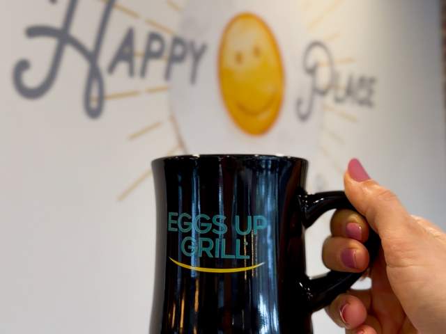 A hand holding a coffee mug in front of a wall with the phrase "this is my happy place" at Eggs Up Grill.