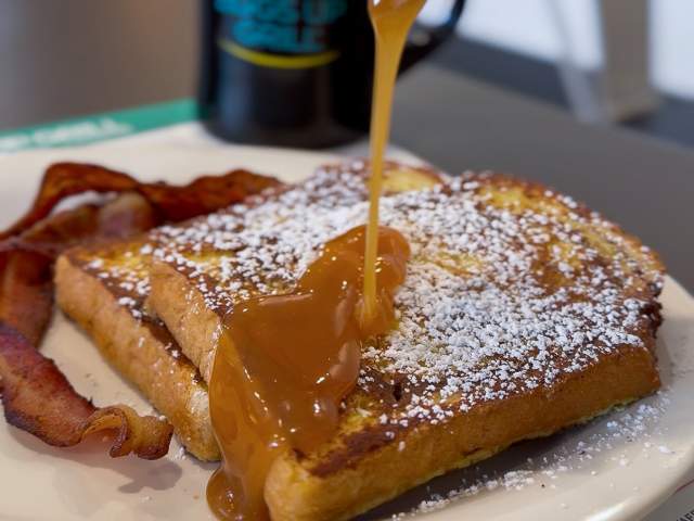 A plate of breakfast food featuring french toast with syrup being poured on it and powdered sugar on top and bacon on the side at Eggs Up Grill.