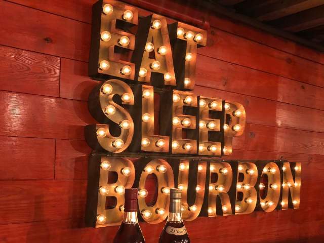 A lightbulb sign that says "Eat, Sleep, Bourbon" with two bottles of alcohol in front of it at Neat Bourbon Bar in downtown Greenville, SC.
