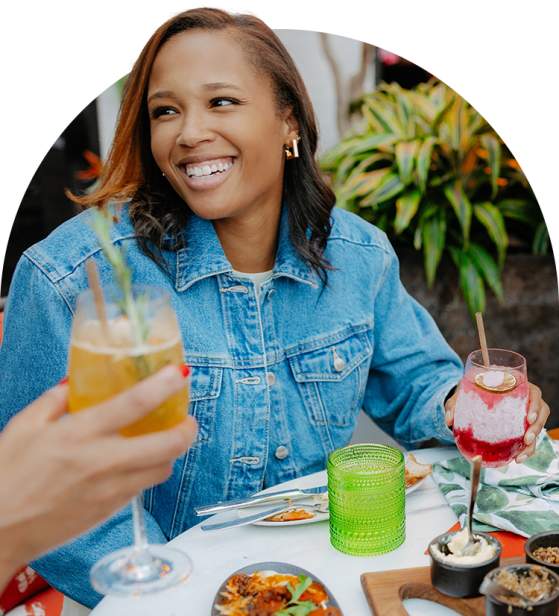 Woman smiling at a table with a delicious food spread and vibrant cocktails.