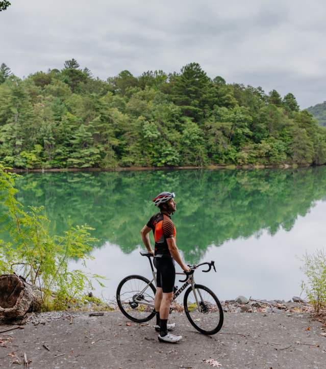 Man wearing helmet and cycling gear with a bike in front of a lake.