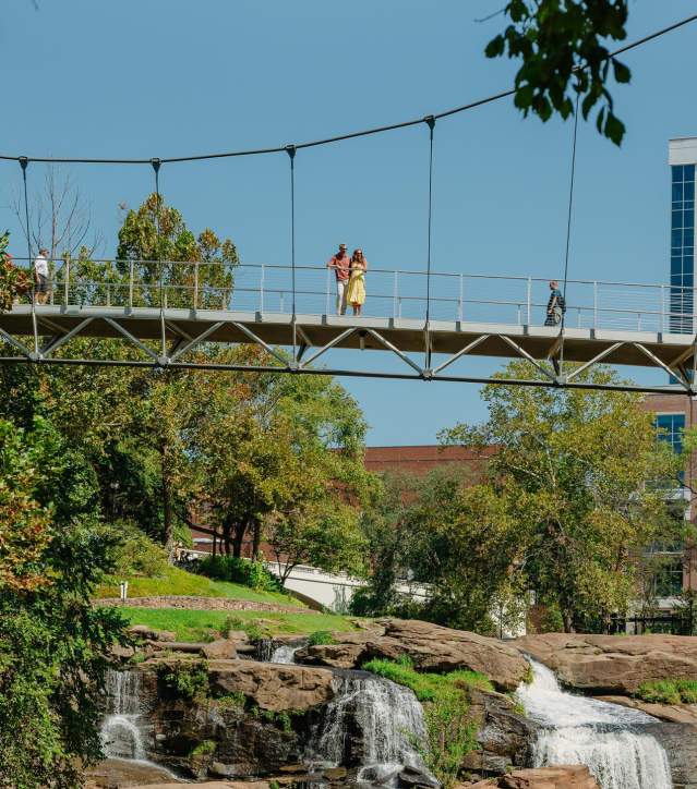 The Liberty Bridge at Falls Park on the Reedy and the view of the waterfalls below and some of the cityscape peeking out from behind the surrounding foliage.