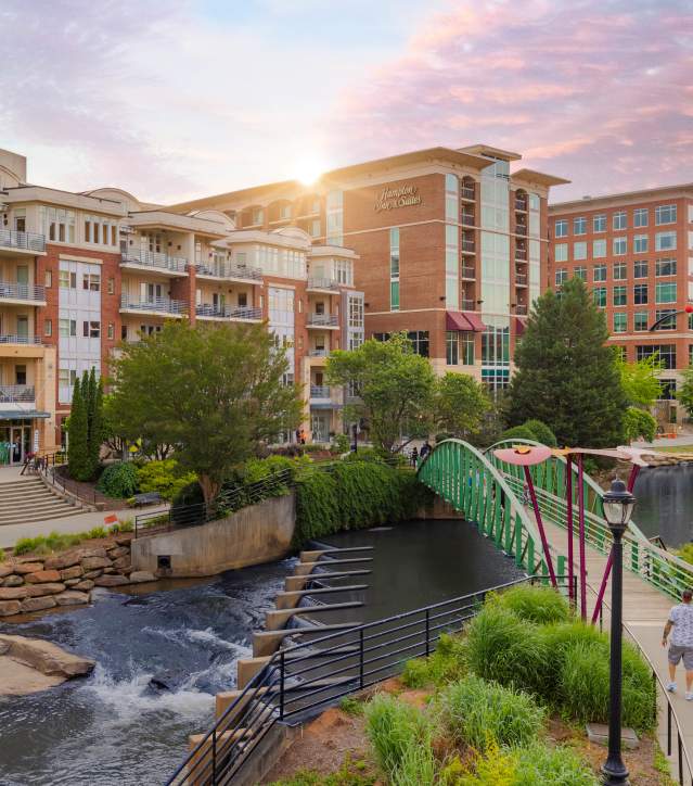 A vibrant sunset over a river, with a green walkable bridge, nearby buildings, and people strolling along the scenic area.