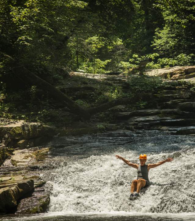 A woman enjoying an adventurous slide down a waterfall, framed by the lush greenery of a forested area.