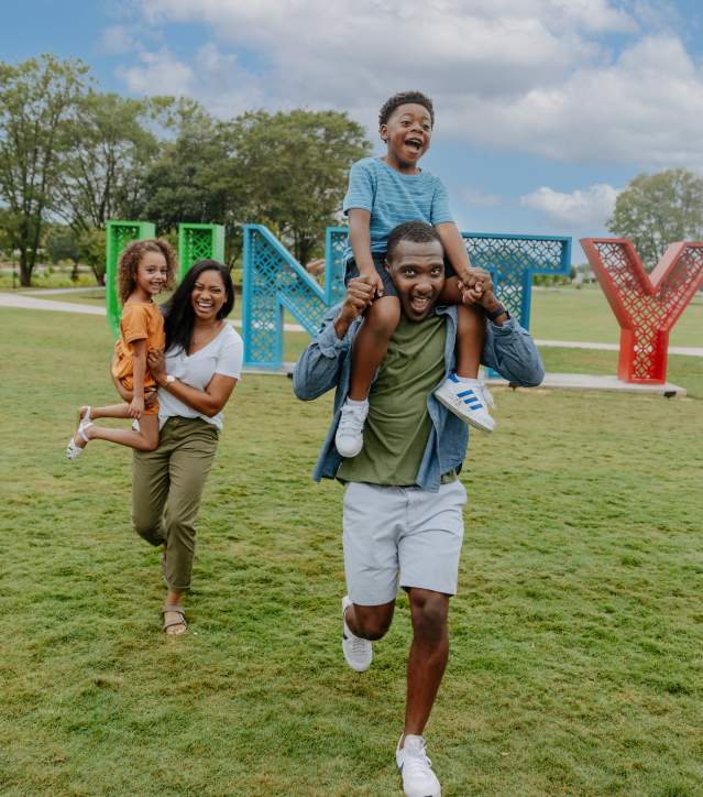 A cheerful family of four playing and running around Unity Park in Greenville, with vibrant greenery and a lively park atmosphere in the background.