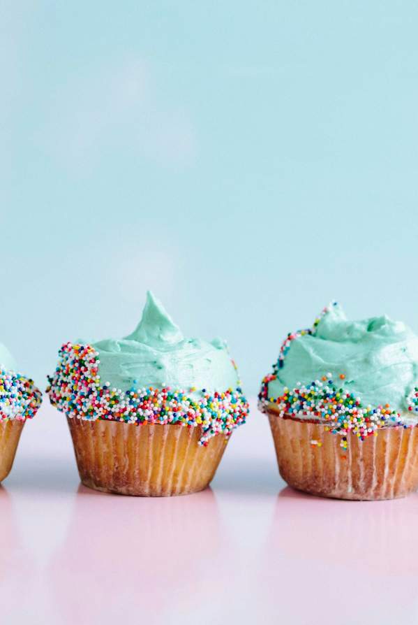 Six cupcakes with teal frosting doused in rainbow sprinkles sit on a pink counter.