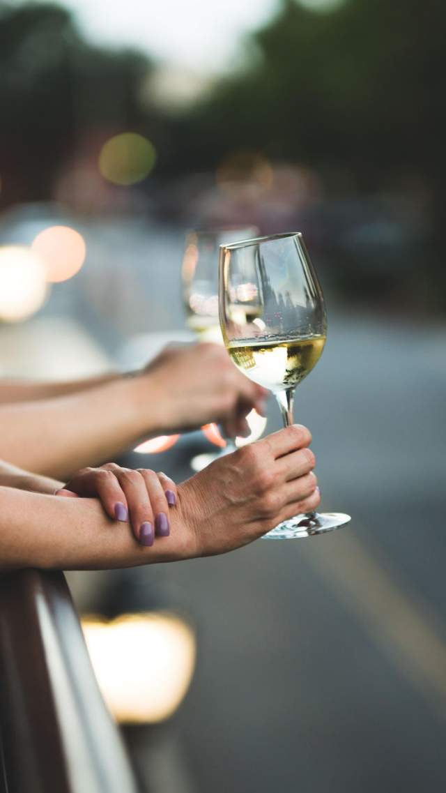 Two glasses of white wine are held out over Jianna's patio with a blurred view of downtown at dusk.