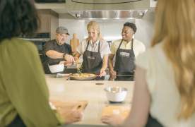 Group of men and women at a cooking class with a chef demonstrating how to prepare a dish on a stove.
