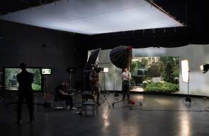 An actor, stage lights, and film crew in front of the LED back wall at Pronk Studios which showcases an LED image of lots of greenery and a small waterfall feature likely at a park.