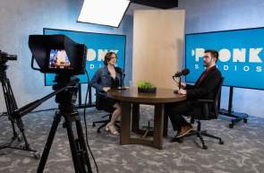 A male and female professionally dressed around a round table with microphones and large monitors behind them with the Pronk Studios logo and film equipment recording their discussion.
