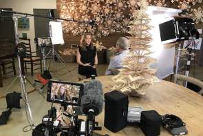 An interview of a man and woman with paper snowflakes cut out and strung in the background and an ornate paper christmas tree made from the paper snowflake cutouts with film equipment surrounding them.