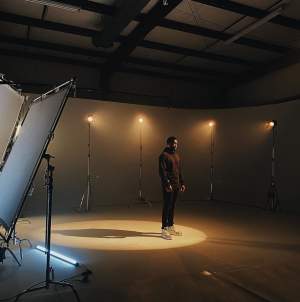 Male subject with yellow tinted, soft spotlight on him casting a shadow in a dark studio with several other yellow, soft lights behind him and film equipment.