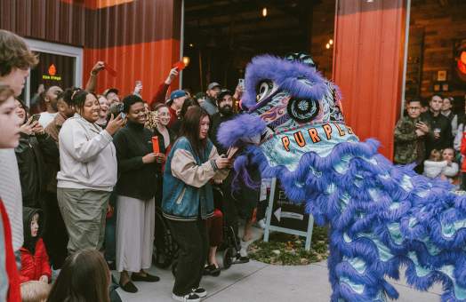 A vibrant purple lion dance costume performs for a crowd of smiling people gathered in front of a red-striped building.