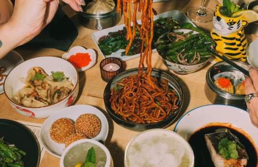 A person uses chopsticks to lift noodles from a bowl on a wooden table filled with various Asian dishes and drinks.