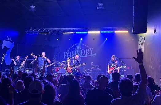 Blue and white stage lights illuminating a stage with five band members on guitars, vocals, and drums at The Foundry at Judson Mill with a crowd in front of them.