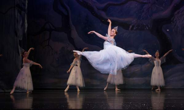 Fair skinned ballerina on stage in a flowy pale blue dress leaping in the air with four ballerinas dancing in the background.