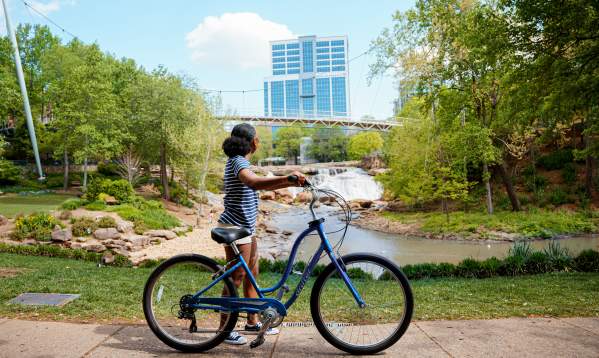 Woman with medium skin tone, blue and white striped shirt, and white shorts standing with a bike admiring the Liberty Bridge at Falls Park on the Reedy in Greenville, SC on a sunny summer day.