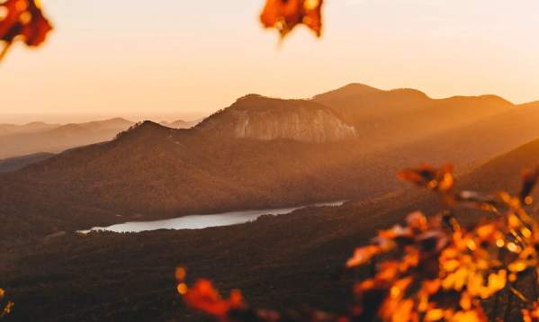 Autumn leaves in the foreground frame a sun-drenched mountain landscape with a lake in the distance.