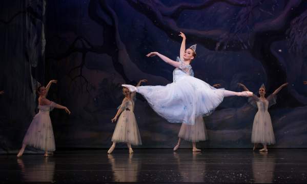Fair skinned ballerina on stage in a flowy pale blue dress leaping in the air with four ballerinas dancing in the background.
