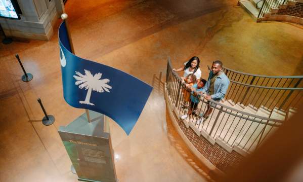 Family of four on a staircase in the Upcountry History Museum gazing up at a sculpture of the South Carolina state flag.
