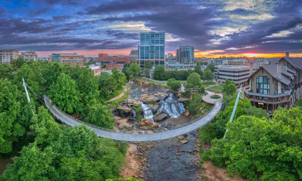 An aerial view of downtown Greenville, featuring the iconic Liberty Bridge spanning over Falls Park, with lush greenery and urban buildings in the background.