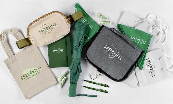 Assortment of new VisitGreenvilleSC branded items, including a mini tote bag, belt bag, umbrella, journal, chapsticks, travel toiletry bag, pet bandana, gift bags, and pens.