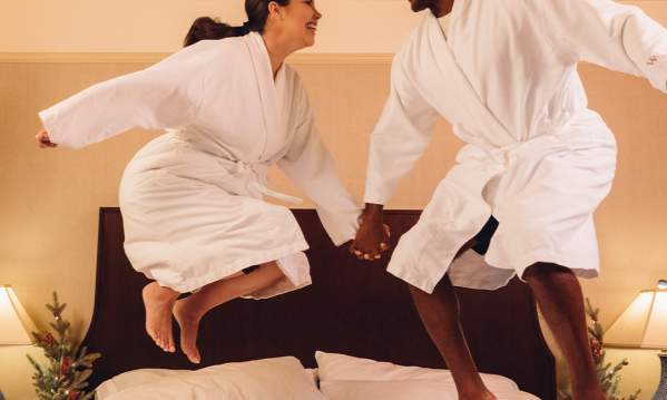 A man and woman holding hands and jumping on a fluffy hotel bed in a hotel suite decorated for Christmas.