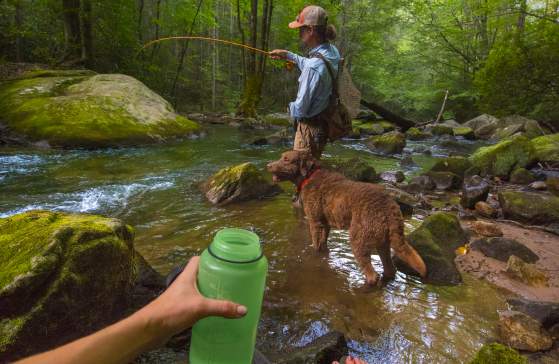 Photo taken from the point of view of a woman wearing leggings and chacos, holding a green water bottle in front of a man fly fishing in a river at Jones Gap with a brown shorthaired dog next to him.