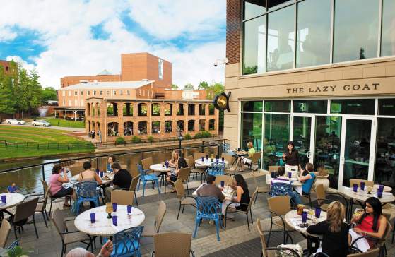 A group of people enjoying a meal on The Lazy Goat’s outdoor patio, surrounded by a vibrant atmosphere and a beautiful view of the Reedy River below.