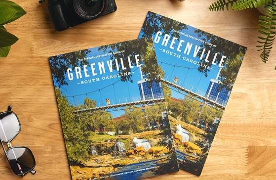 Two copies of the 2025 Official Destination Guide of Greenville, SC, sit on a counter next to a camera, sunglasses, coffee, and a plant.