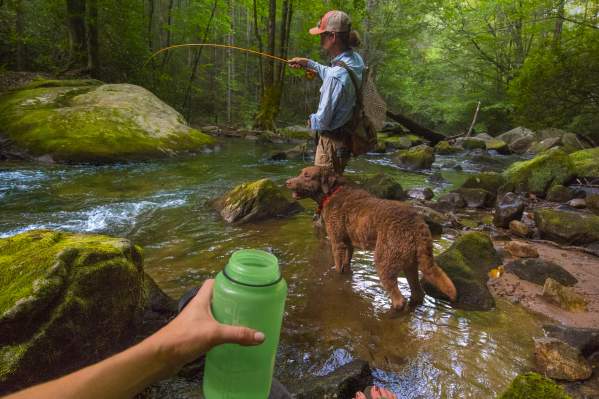 Photo taken from the point of view of a woman wearing leggings and chacos, holding a green water bottle in front of a man fly fishing in a river at Jones Gap with a brown shorthaired dog next to him.
