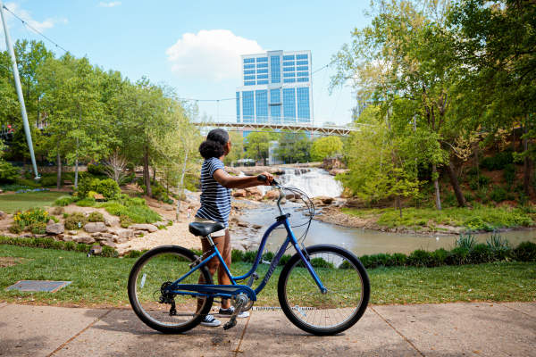 Woman with medium skin tone, blue and white striped shirt, and white shorts standing with a bike admiring the Liberty Bridge at Falls Park on the Reedy in Greenville, SC on a sunny summer day.