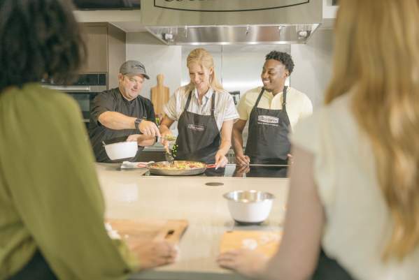 Group of men and women at a cooking class with a chef demonstrating how to prepare a dish on a stove.