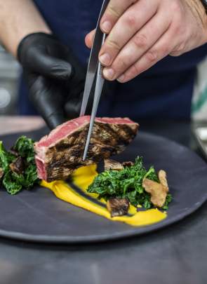 A chef uses tweezers to place a piece of grilled steak onto a black plate with greens and sauce, set against a blurred kitchen background.