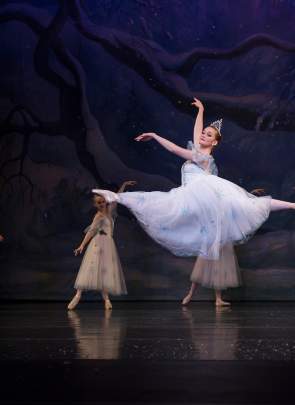 Fair skinned ballerina on stage in a flowy pale blue dress leaping in the air with four ballerinas dancing in the background.
