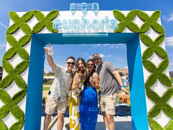 Group of four fair skinned men and women smiling and posing on a sunny day in front of the euphoria photo booth square with greenery and a neon blue euphoria sign that lights up.