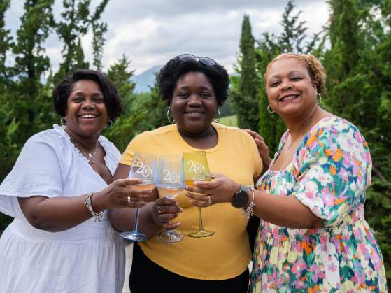 Three smiling women in the foreground hold up wine glasses, with lush green trees and a cloudy sky in the background.
