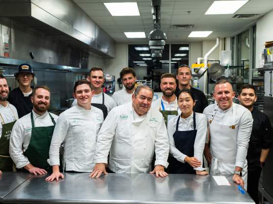 A group of chefs in uniform stand in a professional kitchen, with stainless steel counters in the foreground and kitchen equipment in the background.